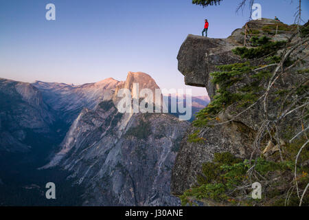 Ein furchtloser Wanderer steht auf einem überhängenden Felsen genießen den Blick in Richtung der berühmten Half Dome am Glacier Point Overlook in Twilight, Yosemite NP, USA Stockfoto