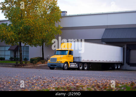 Durchschnittliche Lasttragfähigkeit von Sattelschlepper mit einer leuchtend gelben Kabine mit Dachspoiler und weißem Trockenwagen-Anhänger im Hafenlager Stockfoto