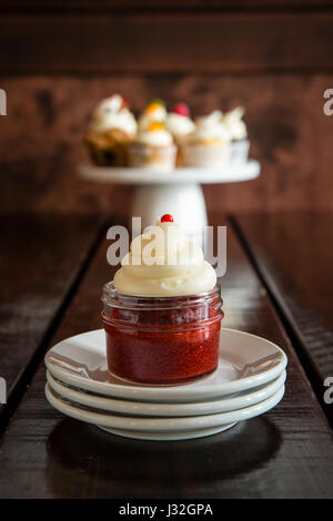 Nahaufnahme von einem rotem Samt Cupcake in einem Glas mit einem Display von Gourmet-Cupcakes im Hintergrund Stockfoto