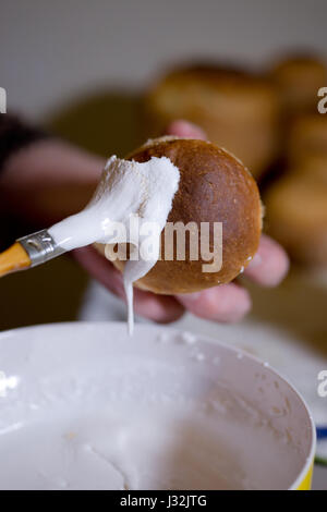 Anwendung der weiße süße Glasur auf die Teig-backen für den Urlaub Tisch mit einem Pinsel, Tauchen Sie ein in die Glasur. Handgemachte Brötchen durch geschickte Gastgeberin. Stockfoto