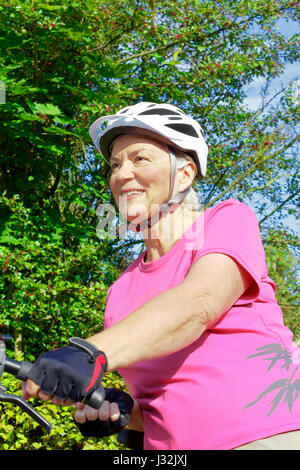 Lächelnde ältere Frau mit weißen Helm und Handschuhe ihr Fahrrad Holding an einem sonnigen Sommer, grüne Büsche im Hintergrund Stockfoto