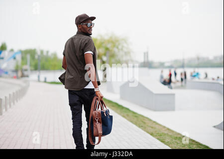Rückansicht-Porträt des Gehens stilvollen afrikanischen amerikanischen Mann Verschleiß an Sonnenbrille und Kappe mit Handtasche im Freien. Streetfashion schwarzen Mann. Stockfoto