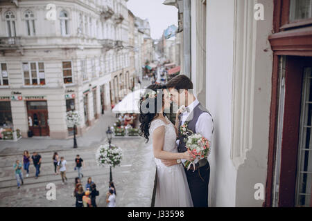 Braut im weißen Kleid stehen auf einem Balkon mit Blick auf die Stadt und Biket, die eine Blume neben ihrem Bräutigam im weißen Hemd Stockfoto