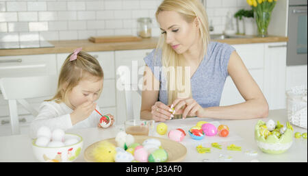 Kleines Mädchen Malvorlagen Ostern Eiern und junge schöne Mutter sitzen in der Nähe und beobachtete in leichte, moderne Küche. Stockfoto