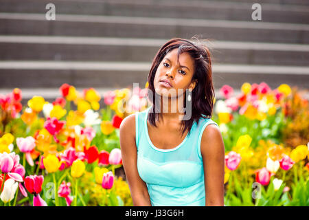 Young African American Woman tragen grüne Tank-Top, Ohrringe, sitzen im Garten mit bunten Blumen, Verkanten Kopf, Blick auf Sie. Stockfoto