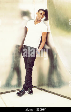 Reflexion. Young African American Mann trägt weiße V Neck T Shirt, schwarze Hose, Lederschuhe, Armbanduhr, Hände in der Tasche, stehen gegen mich zurück Stockfoto