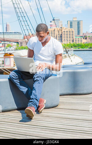 African American Jüngling in New York City Reisen, tragen weiße T Hemd, blaue Hosen, Stiefel Schuhe, Fluss, sitzen auf Laptop-Computer arbeiten. Brookly Stockfoto