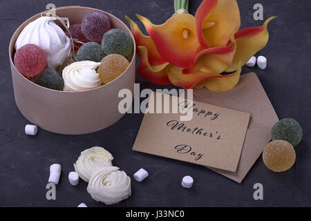 Bouquet von roten gelben Callas mit Marshmallows Marmelade in eine Runde Holzbox und Umschlag mit Beschriftung Muttertag auf grauem Beton Hinterg Stockfoto
