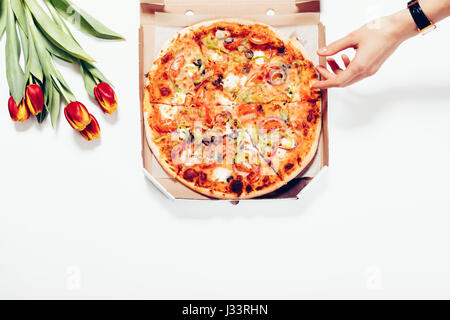 Draufsicht auf eine Pizza in einer Box, Tulpen und eine weibliche Hand auf einem weißen Hintergrund. Das Mädchen nimmt ein Stück Pizza. Stockfoto