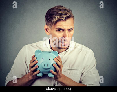 Junge gierige geizig Geschäftsmann hält Sparschwein auf graue Wand Hintergrund isoliert Stockfoto