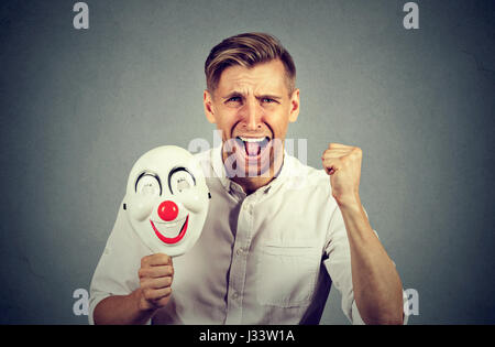 Porträt-junge verärgert böse schreienden Mann hält eine Clownsmaske mit dem Ausdruck ihrer Fröhlichkeit Glück auf graue Wand Hintergrund isoliert. Menschliche Emotionen ja Stockfoto