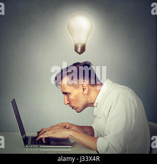 Seite Profil junger Mann arbeiten am Computer sitzen am Schreibtisch mit Glühbirne über dem Kopf auf graue Wand Hintergrund isoliert. Stockfoto