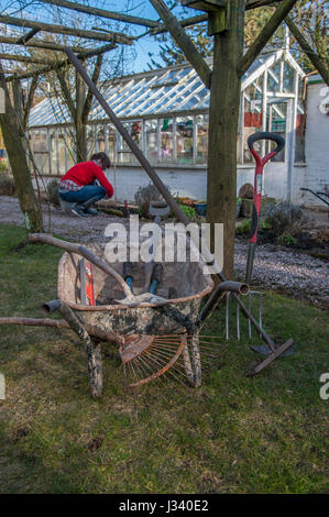 Schubkarre mit Garten-Tools und eine Frau im Hintergrund neben einem Gewächshaus, in einem Garten, Gartenarbeit, Chipping, Lancashire. Stockfoto