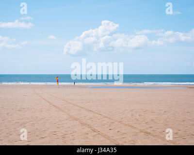 Mann zu Fuß in Richtung Beachflags und die Brandung am Strand von Croyde an einem sonnigen Tag mit blauem Himmel, Devon, England, UK Stockfoto