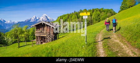 Panoramablick auf der idyllischen Berglandschaft der Alpen mit Wanderer auf Wanderweg entlang frische grüne Almen, Blumen und altes traditionelles Stockfoto