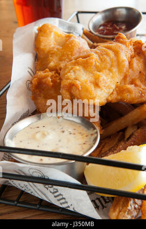 Nahaufnahme eines Korbs von Fish &amp; Chips oder Pommes frites serviert mit Remoulade, Ketchup und ein kühles Bier Stockfoto