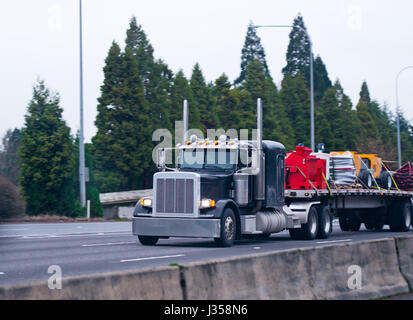 Große klassische amerikanische Leistungsstarke bonnetedbig rig Semi Truck für Langstrecke mit einem flachen Anhänger, wo durch Spanngurte nacheinander Industrielle befestigt sind Stockfoto