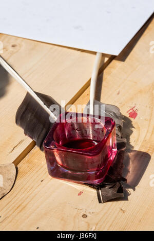 Tintenfaß mit roter Tinte im Inneren Stand über zwei Gans Federn. Blatt Papier in den Hintergrund. Sonnendurchflutetes Holztisch. Niemand um ihn herum. Stockfoto