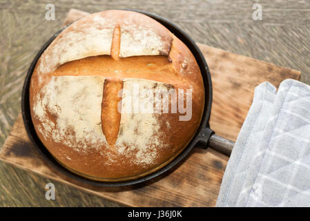 Frisch gebackenes Handwerker Brot in gusseisernen Pfanne auf Holzbrett. Graue Topflappen an Seite. Stockfoto