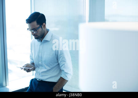 Junger Geschäftsmann mit Mobiltelefon und stützte sich auf Glasfenster im Büro Stockfoto