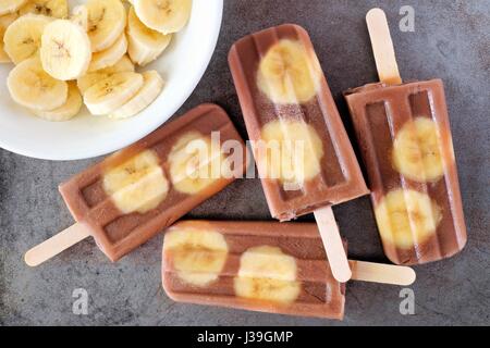Schoko-Bananen Eis am Stiel in einem Cluster mit in Scheiben geschnittenen Bananen auf rustikalen grauen Hintergrund Stockfoto