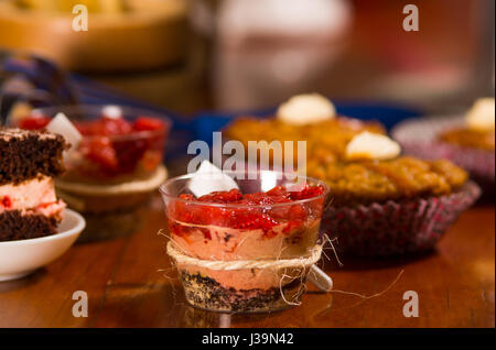 Köstliche Mousse abgedeckt in Erdbeere topping, in kleine Gläser mit Löffeln befestigt, Stücke Kuchen und Muffins Hintergrund, Gebäck-Konzept vorgestellt Stockfoto