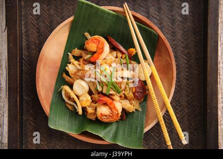 Char Kway Teow. Flache Reisnudeln mit Garnelen, Wurst, Sojabohnensprossen, Ei und Soja-Sauce gebraten. Auf einer hölzernen Platte ausgekleidet mit Bananenblatt serviert. Stockfoto