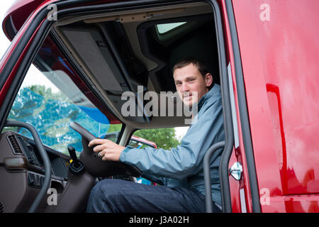 Glücklich LKW-Fahrer in der Kabine des roten LKW hinter dem Lenkrad sitzen und durch die offene Tür suchen. Die moderne Sattelschlepper befindet sich auf dem Parkplatz Stockfoto