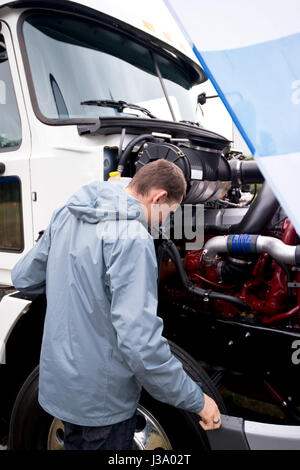 LKW-Fahrer in einer grauen Jacke mit Haube Überprüfung der Betriebsstatus der Komponenten des Motors unter offener Haube von weißen modernen schweren Sattelschlepper Stockfoto