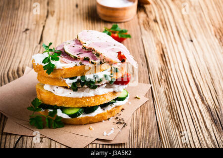 Stapel von Brötchen mit Frischkäse, Schinken, Gurke, Tomate und Frühlingszwiebeln auf rustikalen Holztisch Stockfoto