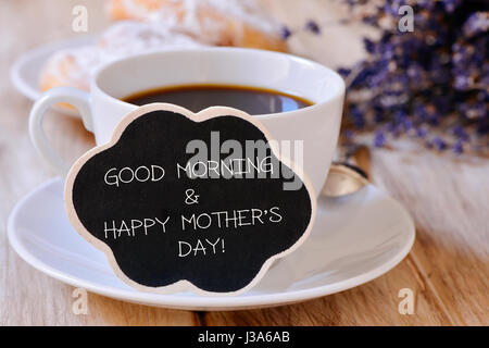 Nahaufnahme der eine schwarze Wolke-förmigen Schild mit dem Text, guten Morgen und einen glücklichen Mütter Tag geschrieben, neben einer Tasse Kaffee auf einem Tisch set für b Stockfoto