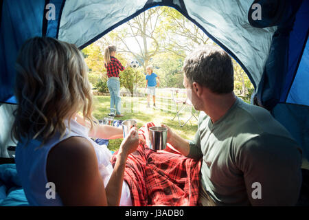 Rückansicht des Paares mit Kaffee und Blick auf Kinder spielen vor dem Zelt Stockfoto