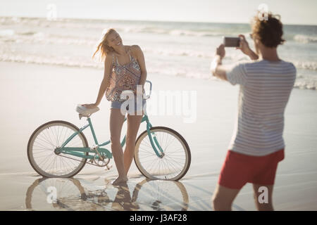 Lächelnde Frau, die mit dem Fahrrad, während Mann Fotografieren sie am Ufer am Strand im sonnigen Tag Stockfoto