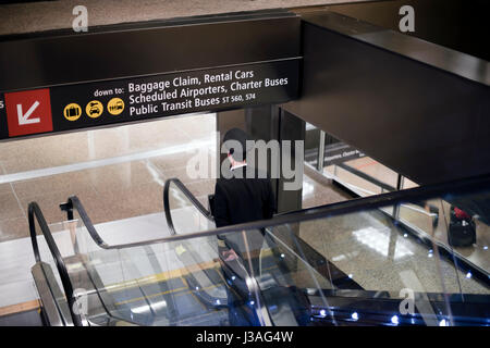 Der Pilot in einem schwarzen Uniform mit Streifen und Kappe mit Koffer in der Hand nach dem Flug auf der Rolltreppe zum Flughafen Erdgeschoss mit Zeichen Stockfoto