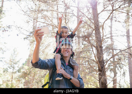 Niedrigen Winkel Ansicht des Vaters Sohn auf Schultern tragen, während der Einnahme von Selfie gegen Bäume im Wald Stockfoto