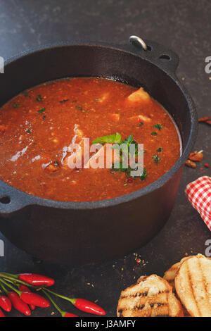 Fisch und Tomaten dünsten in Gusseisen Topf mit gegrillten Brotscheiben auf rustikalen Tisch Makro selektiven Fokus Stockfoto