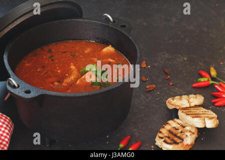 Fisch und Tomaten dünsten in Gusseisen Topf mit gegrillten Brotscheiben auf rustikalen Tisch Makro selektiven Fokus Stockfoto