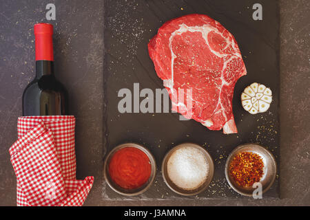 Rohes Fleisch Rib-Eye Steak mit Flasche Rotwein und Gewürzen über Steintisch.  Draufsicht, getönten Jahrgang Bild Stockfoto