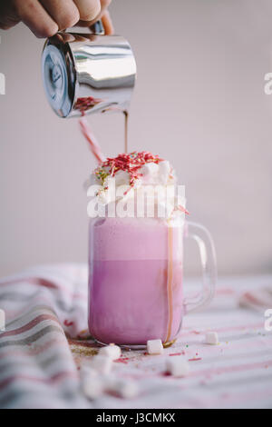 Frau gießt Kaffee in der Tasse farbige Milch mit Sahne, Eibisch und bunte Dekoration. Milch-Shake, Cocktaill, Frappuccino.  Einhorn-Kaffee, u Stockfoto