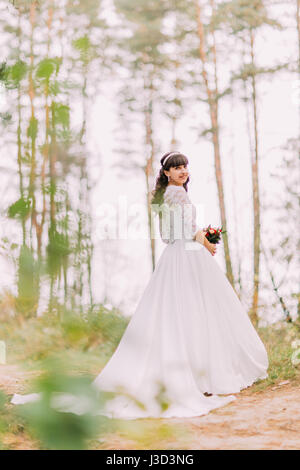 Schöne unschuldige junge Brünette Braut im wunderschönen weißen Kleid steht auf Waldweg am sonnigen Tag Stockfoto