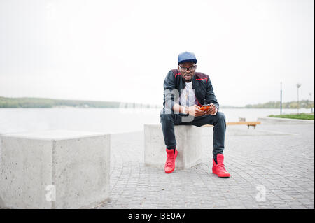 Porträt von stilvollen afrikanischen amerikanischen Mann auf Sportbekleidung, Mütze und Brille sitzt am Steinwürfel mit Telefon zur hand. Schwarze Männer Modell Streetfashion. Stockfoto