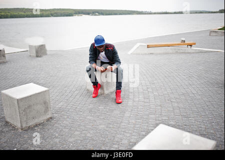 Porträt von stilvollen afrikanischen amerikanischen Mann auf Sportbekleidung, Mütze und Brille sitzt am Steinwürfel mit Telefon zur hand. Schwarze Männer Modell Streetfashion. Stockfoto