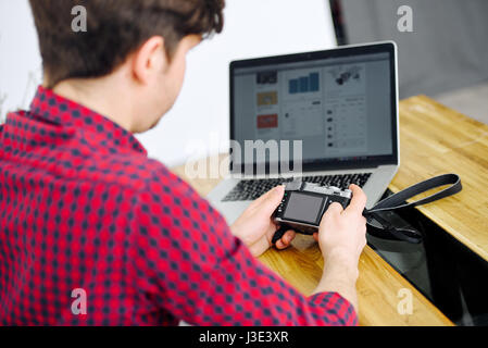 Jungen und erfolgreichen Mann nutzt spiegellose Kamera und sitzt an einem Tisch mit einem Laptop im Büro Stockfoto