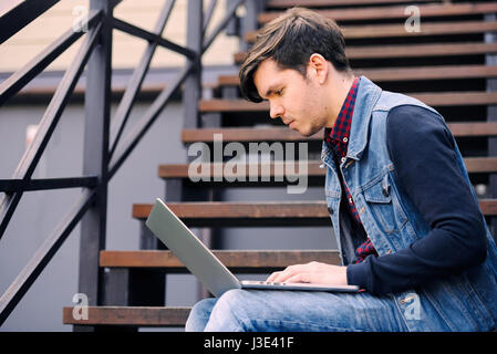 Junger Kerl auf der Straße auf der Treppe sitzt und einen Laptop nutzt. Das Konzept der Lifestyle und Technologie Stockfoto
