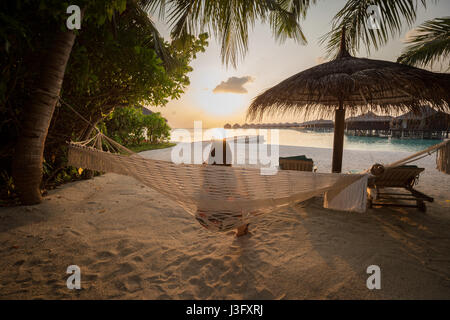 Tropischen Paradies Sonnenuntergang Sonnenaufgang Blick von über Wasserbungalows mit Orange Red Sky sitzen auf Hängematte Stockfoto