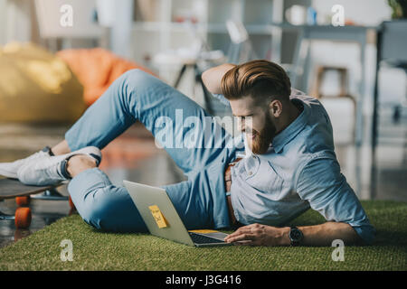 Bärtigen Mann am Boden liegend und mit Laptop in Kreativbüro lächelnd Stockfoto