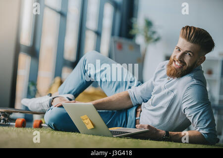 Bärtigen Mann am Boden liegend und mit Laptop in Kreativbüro lächelnd Stockfoto