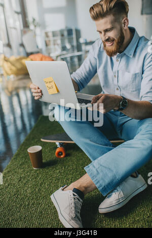 Lächelnd bärtigen Mann sitzen auf Skateboard und Laptop verwenden Stockfoto