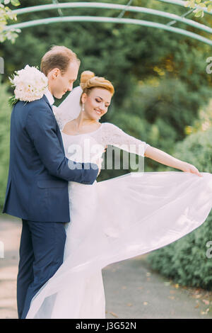 Glückliche junge sanfte Bräutigam und Braut posiert im Park. Braut Hochzeit Blumenstrauß in Händen Stockfoto