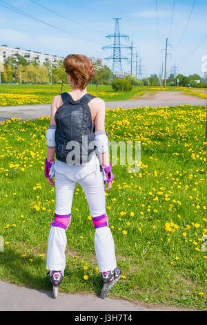 Aktive junge Frau in Sportkleidung auf Rollschuhen. Mit Rücken stehen und schauen in die Ferne. Gegen die hellen grünen Wiese mit Blumen und fernen cit Stockfoto